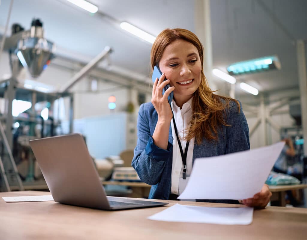 Happy businesswoman communicating over mobile phone while analyzing reports in a factory.